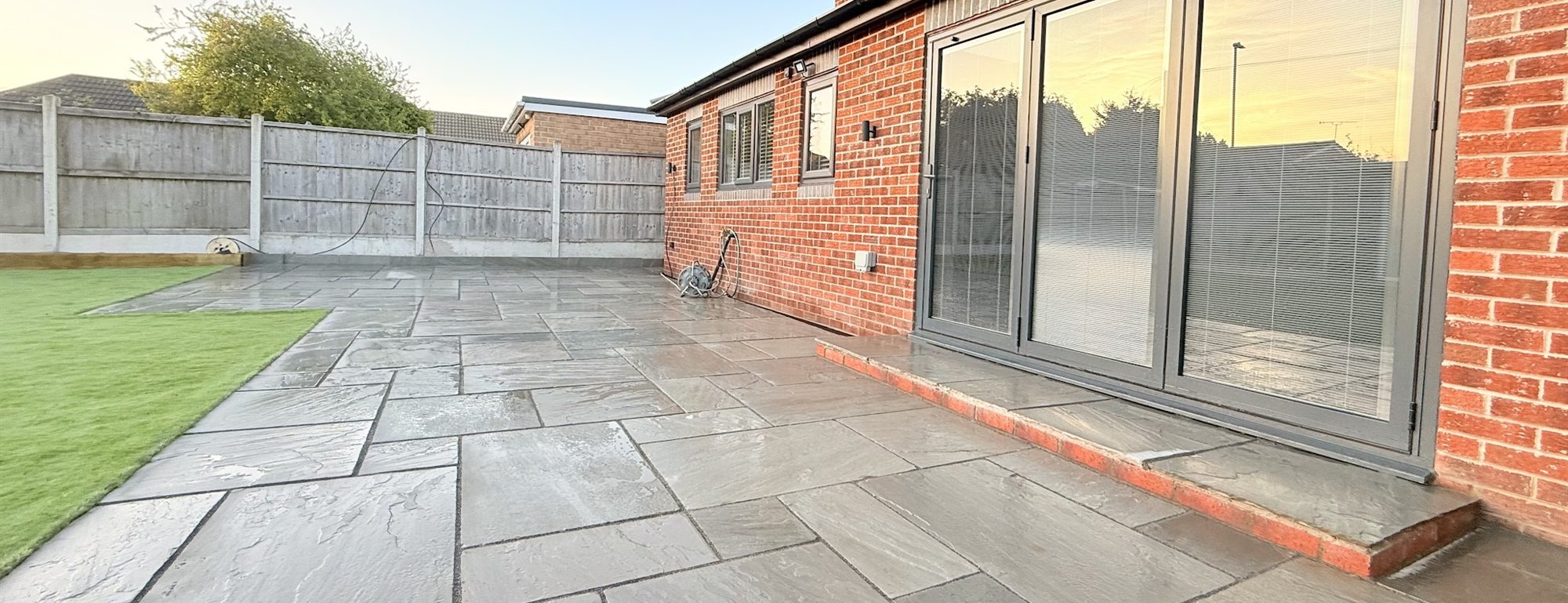 Wide Indian sandstone patio laid across a rear garden beside a brick house at dusk.