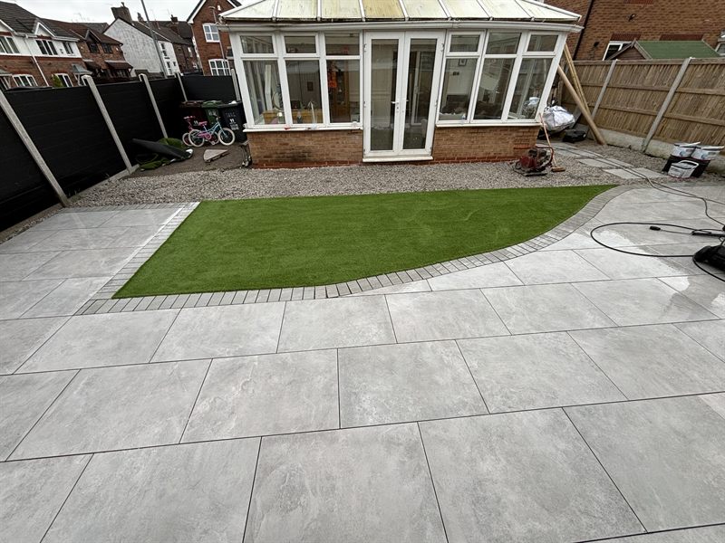 Pale patio curving around an artificial grass lawn beside a conservatory.