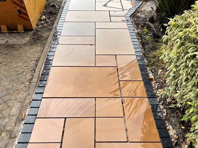 Buff Indian sandstone path edged with charcoal setts, wet from rain.
