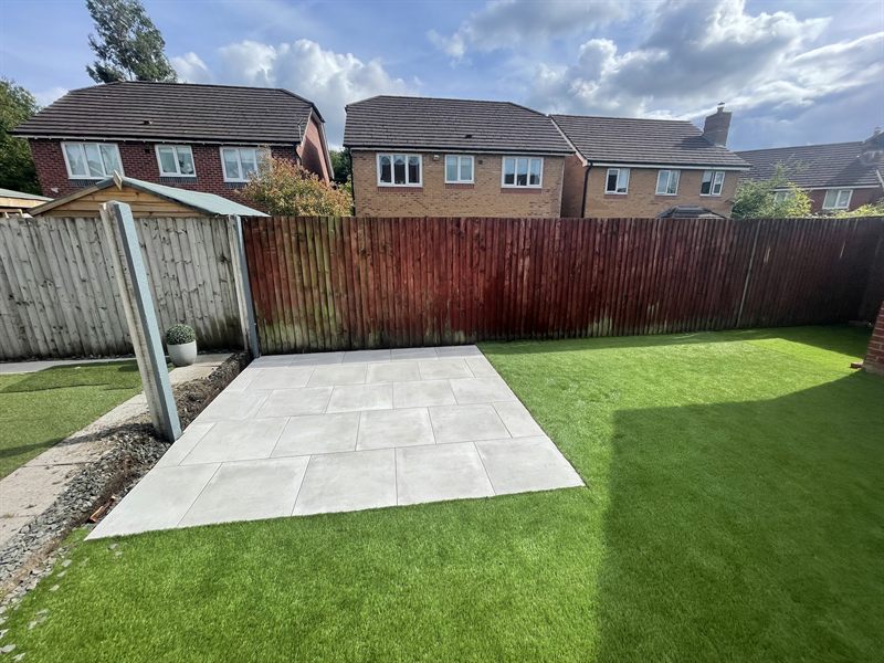 Artificial grass either side of a pale patio square in a fenced garden.