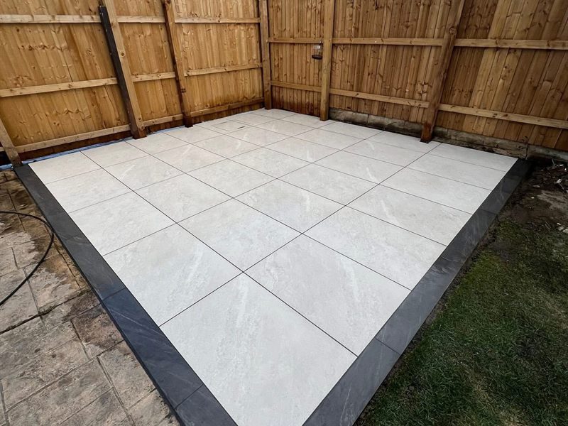 Pale porcelain patio with a charcoal border laid square into a fenced corner.