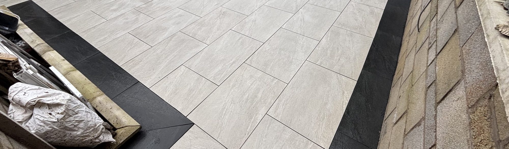 Pale grey porcelain patio with a dark border, laid against a stone retaining wall.
