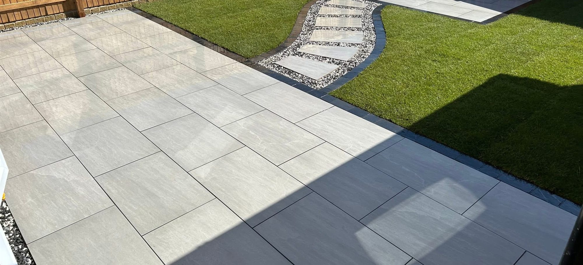 Large grey porcelain patio with a stepping-stone path curving through a lawn, edged with dark trim.