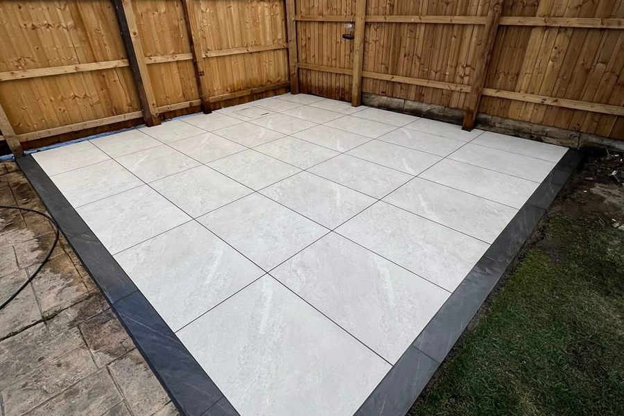 Pale porcelain patio with a charcoal border, laid square into a fenced garden corner.